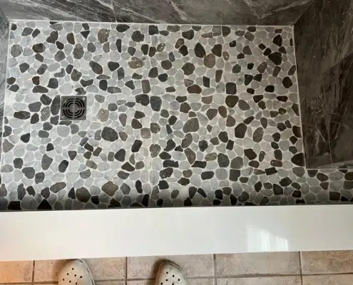Sliced river rock mosaic shower floor in shades of charcoal and tan with a square stainless steel drain, showcasing a slip-resistant and organic design for a custom NH bathroom renovation