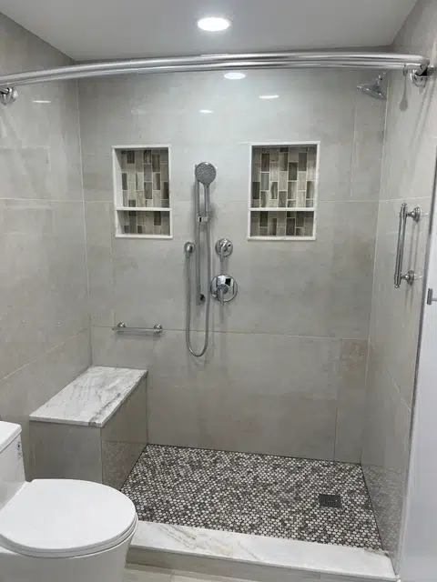 Large-format grey tiled shower walls installed over a professional waterproof backing system, featuring double built-in niches and pebble flooring.