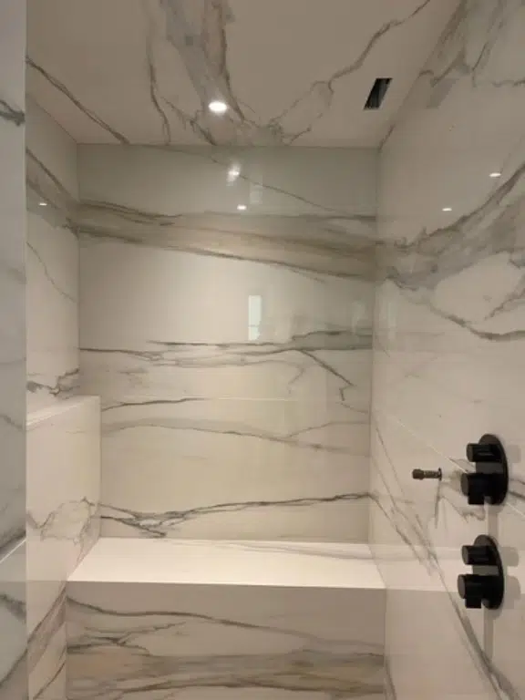 Luxurious seamless shower showing solid quartz slabs with continuous marble veining running floor-to-ceiling, a matching bench, and black fixtures.