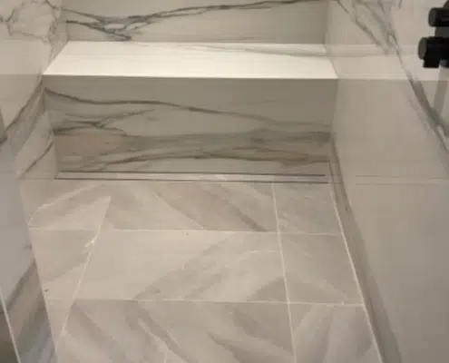 Modern bathroom remodel showing a luxury shower with marble-look slabs, a built-in floating bench, matte black fixtures, and a linear drain.
