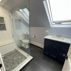 Clear frameless glass shower enclosure with a pebble stone floor and tiled niche under a skylight, beside a navy vanity in a small bathroom.
