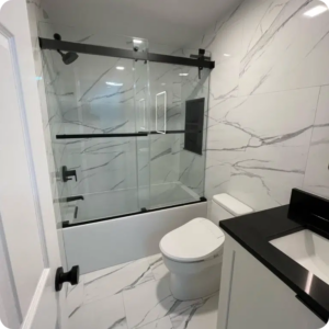 Large-format marble-look porcelain tile runs floor to ceiling around a tub with a black sliding glass door in a small bathroom remodel.