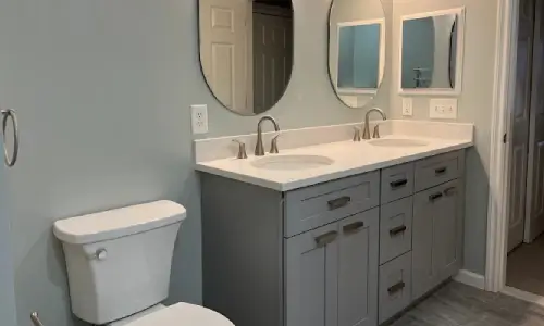 Double vanity with gray shaker cabinets, a white top and two oval mirrors fits beside the toilet in a compact bathroom, a common New Hampshire layout.
