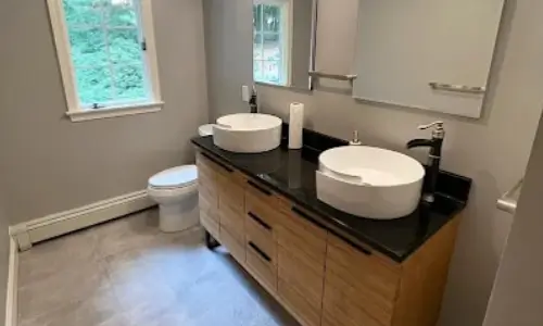 Large gray floor tiles, wide mirrors and a window keep this bathroom feeling open around a wood vanity with a black top and two round vessel sinks.